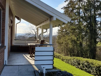 Eingang Ferienwohnung Schulz Überdachte Terrasse einer Ferienwohnung mit Holzmöbeln, umgeben von grünen Bäumen und Rasen.