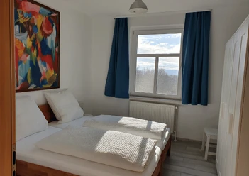 Schlafzimmer Ferienwohnung Schulz Helles Schlafzimmer mit Doppelbett, bunter Wandkunst, Fensteraussicht auf Berge und weiße Möbel.