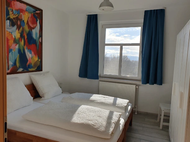 Schlafzimmer Ferienwohnung Schulz Helles Schlafzimmer mit Doppelbett, bunter Wandkunst, Fensteraussicht auf Berge und weiße Möbel.