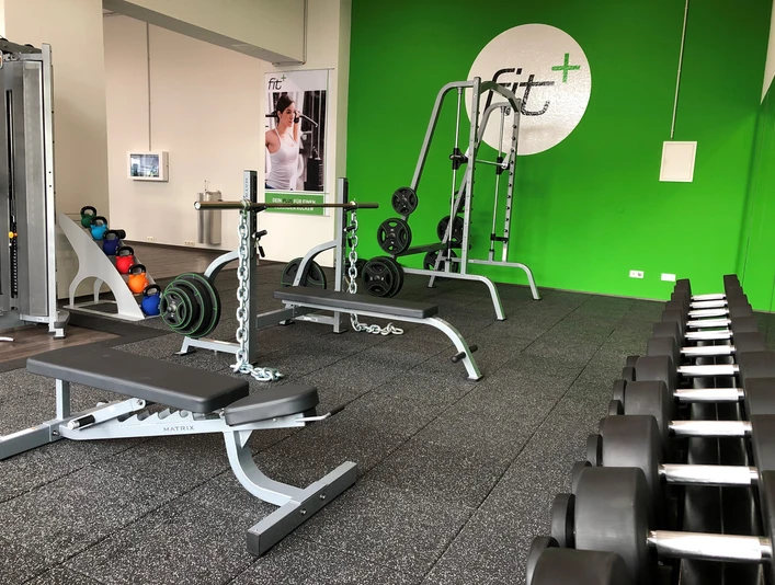 fitplus-wiefelstede-freihantel Freihantelbereich im modernen Fitnessstudio: Bänke, Hanteln und ein heller, grüner Raum.