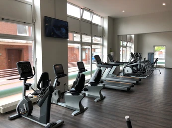 fitplus-wiefelstede-cardio Ein heller, moderner Fitnessraum mit einer Reihe von Cardiogeräten. Große Fenster sorgen für viel Licht.