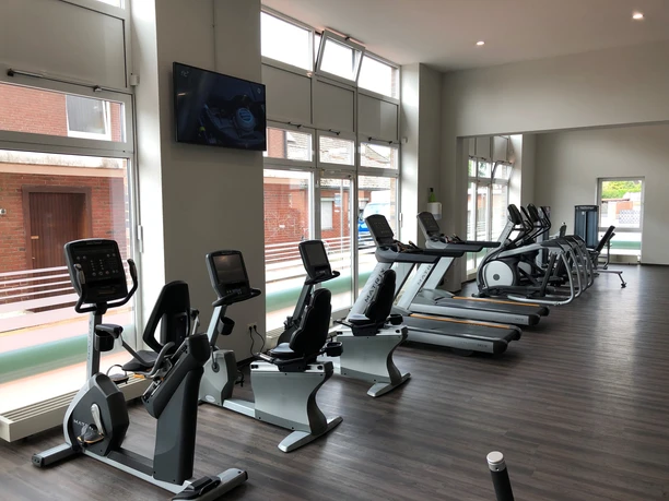 fitplus-wiefelstede-cardio Ein heller, moderner Fitnessraum mit einer Reihe von Cardiogeräten. Große Fenster sorgen für viel Licht.