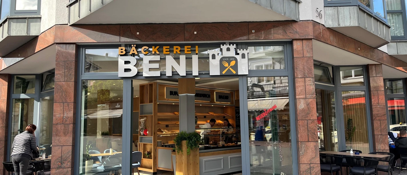 Eingang der Bäckerei Beni mit großer Glasfront, Auslage und Sitzplätzen im Freien