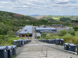 :metabolon Stufen führen hinunter zu einem Gebäude, flankiert von Mülltonnen, umgeben von grüner Landschaft.