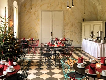 Gemütlicher Raum mit Weihnachtsbaum, runden Tischen und roten Servietten, gedeckt mit Tassen und Kerzen; im Hintergrund eine Anrichte mit Getränken.Cozy room with Christmas tree, round tables and red napkins, set with cups and candles; in the background a sideboard with drinks.Útulná místnost s vánočním stromkem, kulatými stoly a červenými ubrousky, prostřenými šálky a svíčkami; v pozadí příborník s nápoji.Przytulny pokój z choinką, okrągłymi stołami i czerwonymi serwetkami, zastawiony filiżankami i świecami; w tle kredens z napojami.Gezellige kamer met een kerstboom, ronde tafels en rode servetten, gedekt met kopjes en kaarsen; een dressoir met drankjes op de achtergrond.Sala accogliente con albero di Natale, tavoli rotondi e tovaglioli rossi, apparecchiati con tazze e candele; sullo sfondo una credenza con bevande.