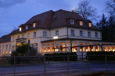 Zeitgeist Sächsisches Haus.JPG Großes, beleuchtetes Gebäude bei Dämmerung, mit roten Vorhängen und Weihnachtsdekoration in den Fenstern, umgeben von Bäumen.