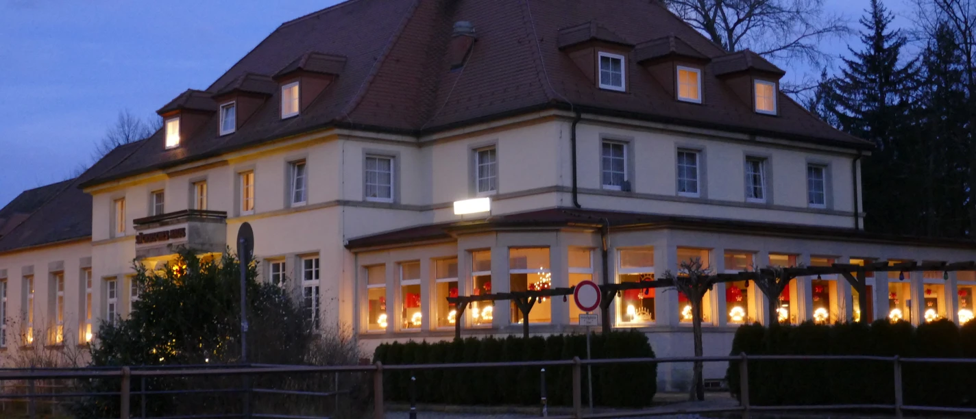 Zeitgeist Sächsisches Haus.JPG Großes, beleuchtetes Gebäude bei Dämmerung, mit roten Vorhängen und Weihnachtsdekoration in den Fenstern, umgeben von Bäumen.