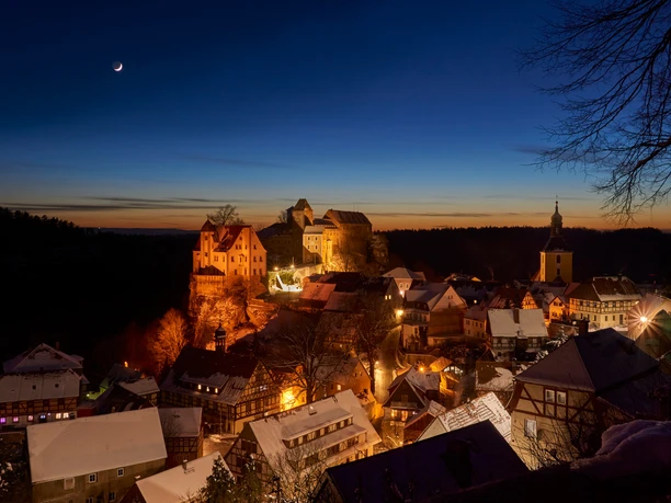 Hohnstein Castle towers over the snow-covered, festively illuminated town in the winter evening light.