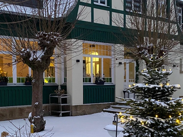 Pohled zvenčí na tradiční restauraci Ottomühle Zasněžená zahrada s osvětleným vánočním stromem před hrázděným domem se zelenými a bílými fasádami, uvnitř teplé osvětlení.