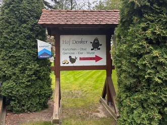 Hofladen Denker Hölzernes Schild des Hofladens Denker mit Hinweis auf Kartoffeln, Gemüse und Eier, umrahmt von Nadelbäumen.
