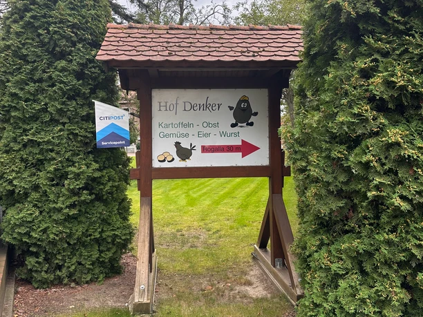 Hofladen Denker Hölzernes Schild des Hofladens Denker mit Hinweis auf Kartoffeln, Gemüse und Eier, umrahmt von Nadelbäumen.