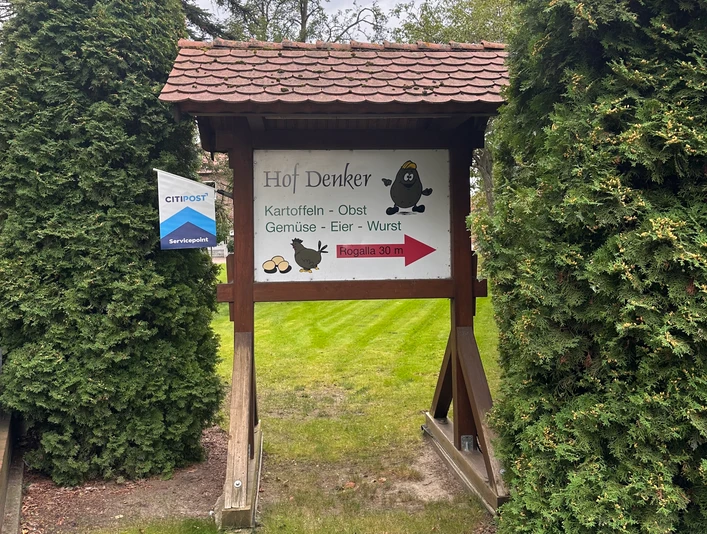 Hofladen Denker Hölzernes Schild des Hofladens Denker mit Hinweis auf Kartoffeln, Gemüse und Eier, umrahmt von Nadelbäumen.