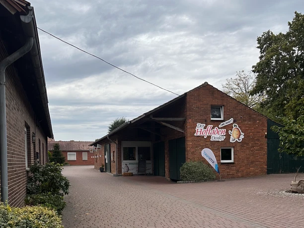 Hofladen Denker Ein Hofladen aus rotem Backstein mit Werbeschildern und einer kleinen Terrasse vor dem Eingang.