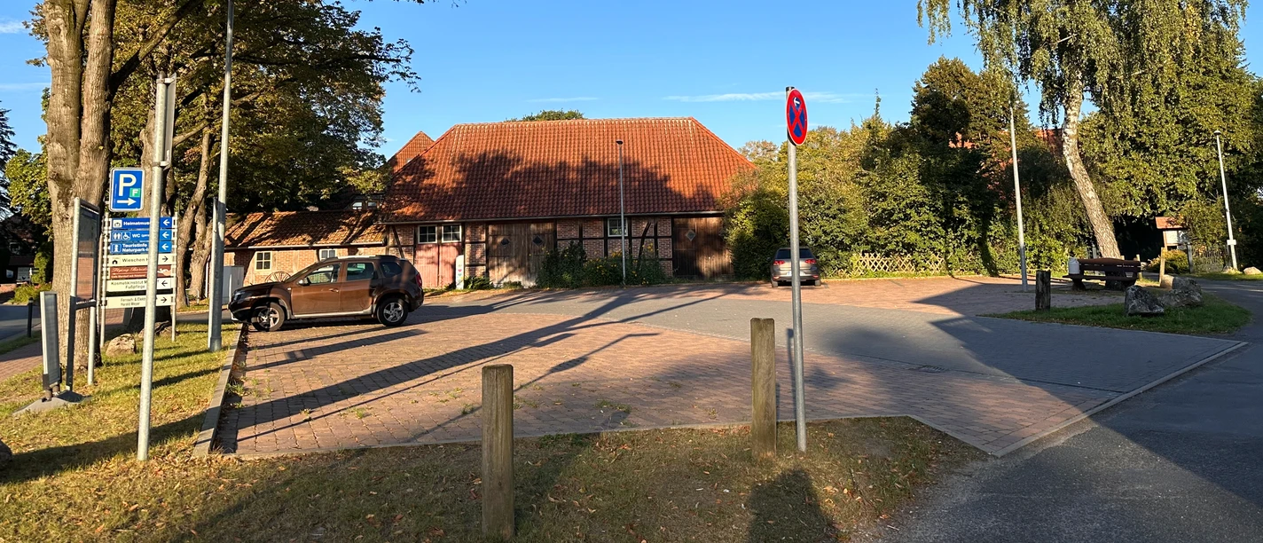 Parkplatz mit Pflastersteinen, umrahmt von Bäumen und einem Gebäude mit rotem Ziegeldach.