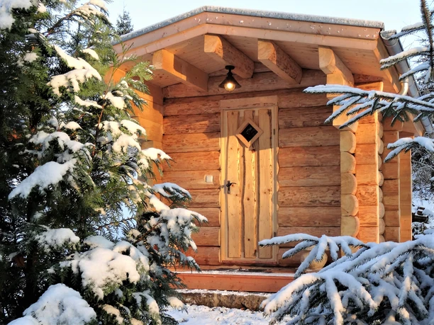 Finnische Blockhaussauna Forsthaus Bielatal Holzhütte im Schnee, umgeben von schneebedeckten Tannen. Eine Lampe leuchtet über der Tür und die Atmosphäre wirkt ruhig und winterlich.