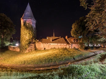 Nachtaufnahme des Wehrturms im Winkel, umgeben von Bäumen, mit beleuchtetem Mauerwerk und Gras.