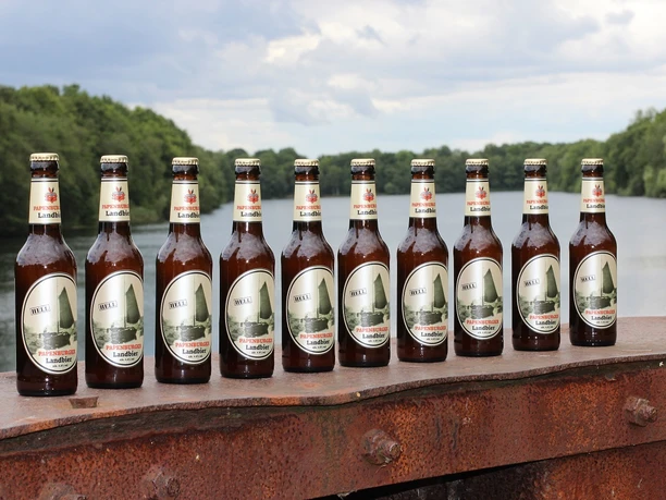 Papenburger Landbier Reihe Papenburger Landbier-Flaschen auf rostigem Geländer vor Fluss und grüner Baumkulisse
