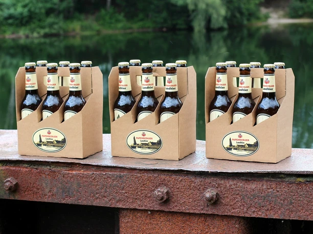 Papenburger Landbier Drei Sixpacks Papenburger Landbier auf rostiger Metallkante vor grünem Seeufer arrangiert.