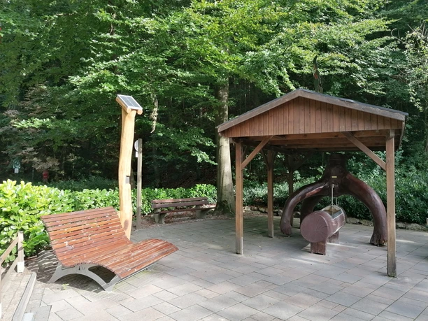 Hörstation Anna & Hermann in Bad Iburg Interaktive Hörstation mit Waldgeräuschen unter Holzpavillon nahe einer Ruhebank in einem bewaldeten Park.