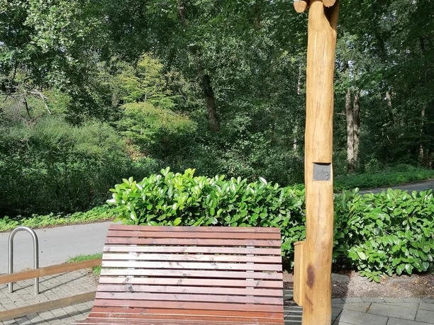 Hörstation Anna & Hermann in Bad Iburg Holzbank mit Rückenlehne neben einer naturholzfarbenen Hörstation in einem Waldbereich.