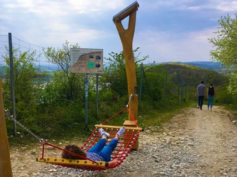 Hörstation 05 in Lengerich Eine Person entspannt sich in einer Hängematte entlang eines Wanderwegs mit grüner Landschaft im Hintergrund.