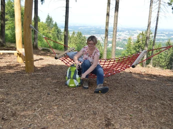 Eine Frau sitzt in einer roten Hängematte im Wald, backpacker-rastend mit Ausblick auf eine Stadt.