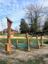 Hörstation Anna & Hermann in Bielefeld Ein Spielplatz mit Kletternetz und Holzgestellen, umgeben von Bäumen und einem gepflasterten Weg.