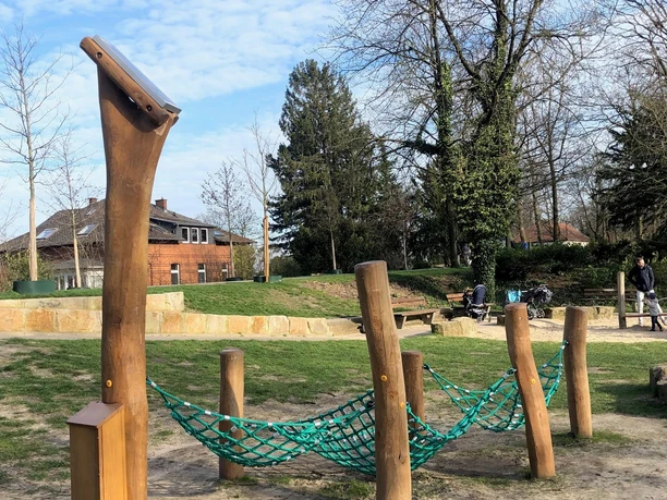 Hörstation Anna & Hermann in Bielefeld Ein Spielplatz mit Kletternetz und Holzgestellen, umgeben von Bäumen und einem gepflasterten Weg.