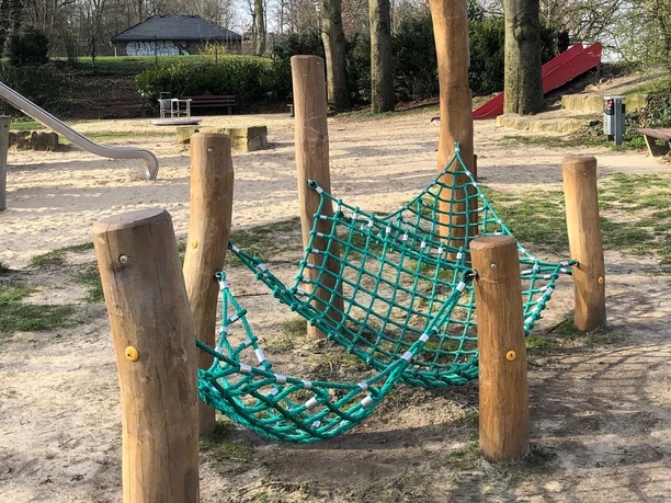 Hörstation Anna & Hermann in Bielefeld Holzpfosten mit grünem Kletternetz auf einem Spielplatz, umgeben von Bäumen und einem Hügel.