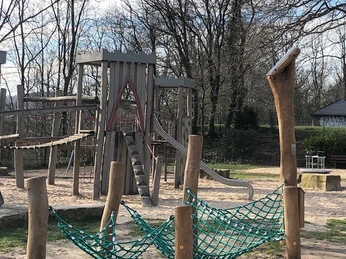 Hörstation Anna & Hermann in Bielefeld Holzspielplatz mit Klettergerüst und Netzschaukel, umgeben von Bäumen und offenem Himmel.
