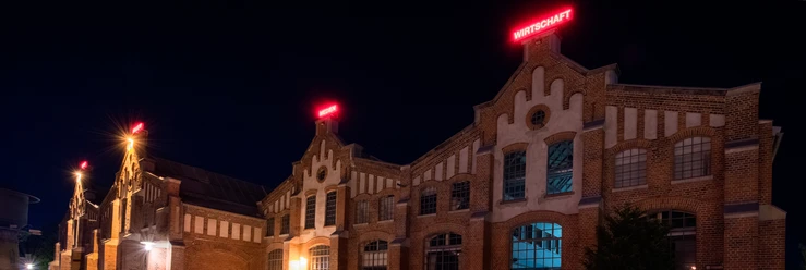 Halle IV Backsteinbau mit großen Fenstern und roter Leuchtschrift, nachts beleuchtete Veranstaltungshalle.