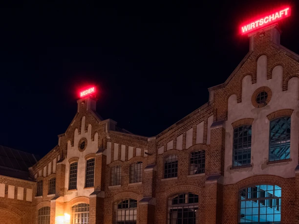 Halle IV Backsteinbau mit großen Fenstern und roter Leuchtschrift, nachts beleuchtete Veranstaltungshalle.