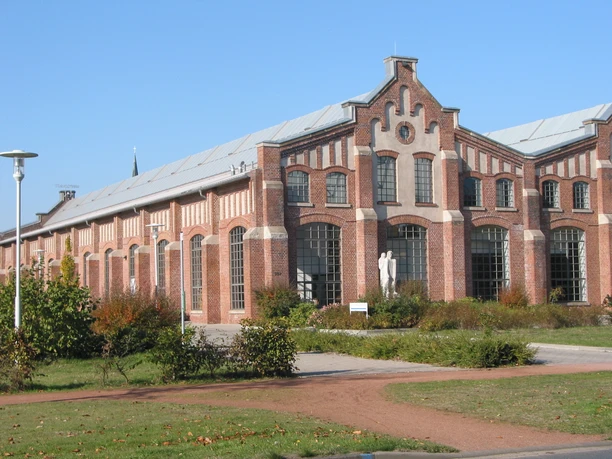 Halle IV Backsteinbau mit großen Rundbogenfenstern, Skulptur im Eingangsbereich und begrüntem Vorplatz.