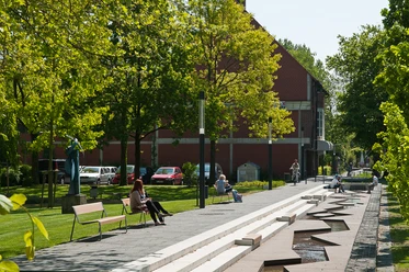 Stadtgrabenpromenade Grüne Promenade mit Wasserlauf, Sitzbänken und Spaziergängern unter Bäumen nahe rotem Ziegelbau.