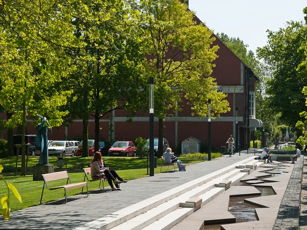 Stadtgrabenpromenade Grüne Promenade mit Wasserlauf, Sitzbänken und Spaziergängern unter Bäumen nahe rotem Ziegelbau.