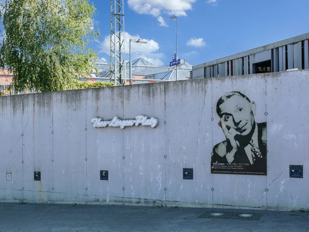 Theo Lingen Graue Betonwand mit Porträt von Theo Lingen und Schriftzug Theo-Lingen-Platz bei blauem Himmel