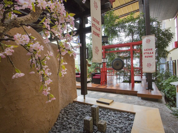 Saunalandschaft - Japanischer Garten Japanischer Garten mit Kirschblüten, Steinen und Gong, umgeben von Bannern mit ruhigen Botschaften.