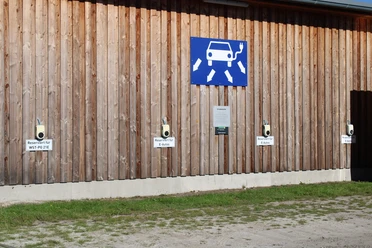 ladestation-park-der-gaerten.jpg Holzwand mit E-Ladesäulen, markiert für „E-Autos“, unter Zeichen für Elektromobilität.