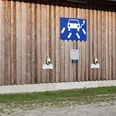 ladestation-park-der-gaerten.jpg Holzwand mit E-Ladesäulen, markiert für „E-Autos“, unter Zeichen für Elektromobilität.