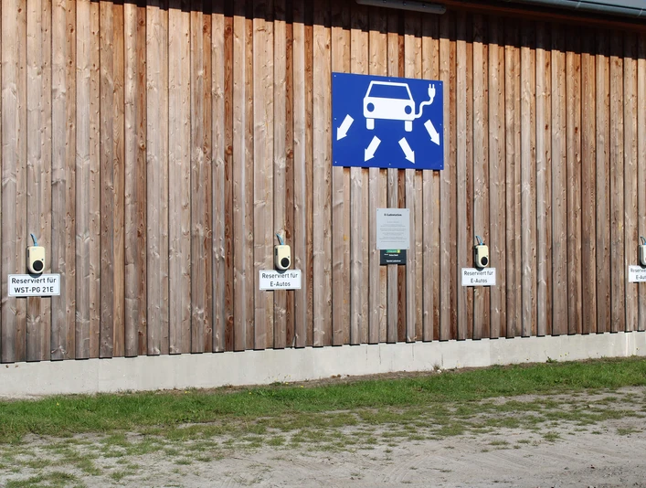 ladestation-park-der-gaerten.jpg Holzwand mit E-Ladesäulen, markiert für „E-Autos“, unter Zeichen für Elektromobilität.