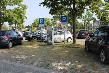 ladestation-sued1-zob.JPG Ein Elektroauto parkt an einer Ladestation zwischen grünen Bäumen und mehreren Fahrzeugen in einer urbanen Umgebung.