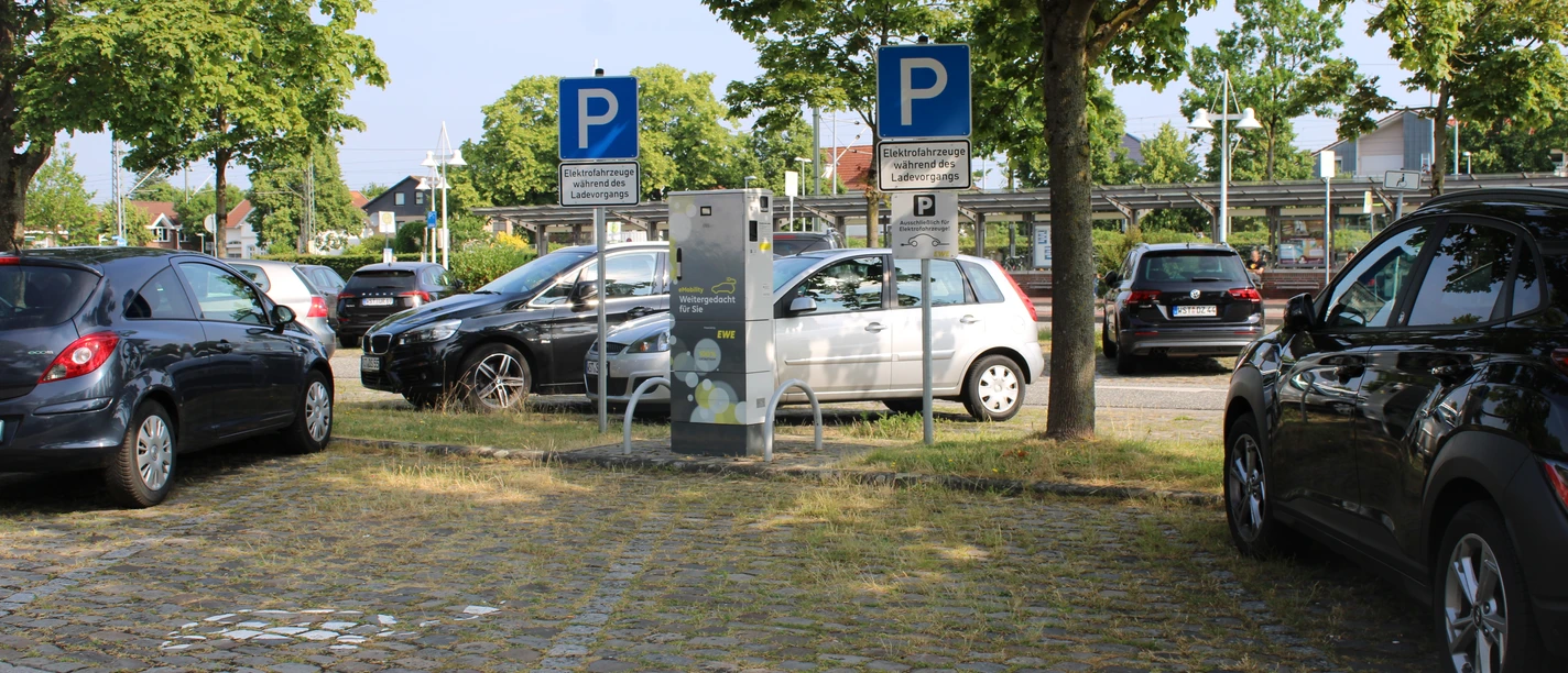 ladestation-sued1-zob.JPG Ein Elektroauto parkt an einer Ladestation zwischen grünen Bäumen und mehreren Fahrzeugen in einer urbanen Umgebung.