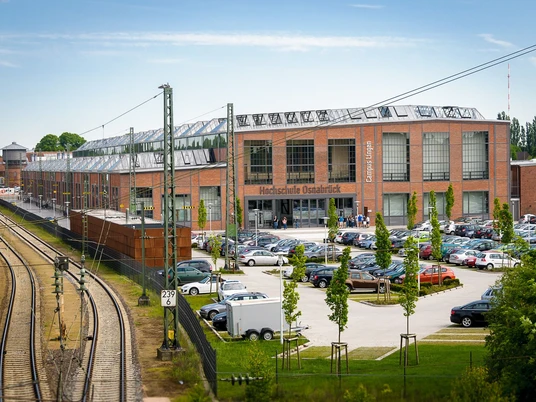 Campus Lingen Backsteingebäude des Campus Lingen mit Glasdach, angrenzenden Bahngleisen und Parkplatz.
