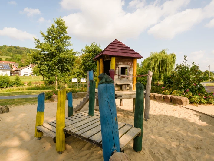 Spielplatz im Emmerauenpark mit Holzturm, Rutsche, bunten Holzpfosten, Sandboden und Bäumen.
