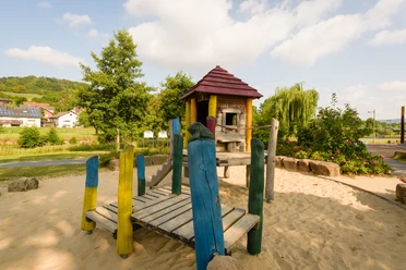 Abenteuerspielplatz im Emmerauenpark Spielplatz im Emmerauenpark mit Holzturm, Rutsche, bunten Holzpfosten, Sandboden und Bäumen.
