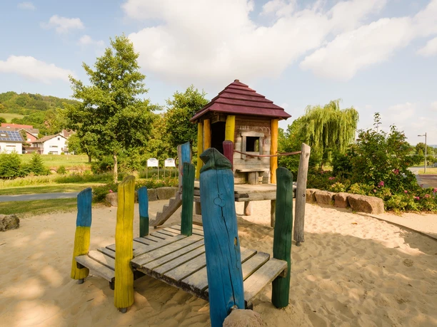 Abenteuerspielplatz im Emmerauenpark Spielplatz im Emmerauenpark mit Holzturm, Rutsche, bunten Holzpfosten, Sandboden und Bäumen.