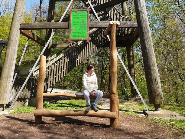 Hörstation am Luisenturm in Borgholzhausen Eine Person sitzt auf einem Holzschaukelstuhl vor dem Luisenturm in einem grünen Waldgebiet in Borgholzhausen.