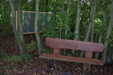 Grenzbank Holzbank mit Inschrift "Grenzbank" steht zwischen schattigen Bäumen, nahe einem Infoboard im Wald.