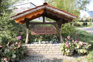 Erholungsort Hummersen Holzbalken-Struktur mit Schild "Erholungsort Hummersen", umgeben von üppigen Pflanzen und Blumen.