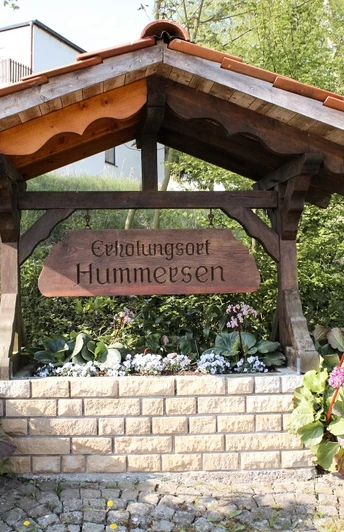 Erholungsort Hummersen Holzbalken-Struktur mit Schild "Erholungsort Hummersen", umgeben von üppigen Pflanzen und Blumen.